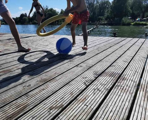 zwei Kinder in Badehosen spielen auf einem Holzsteg am See mit einem Wasserball und einer Schwimmnudel