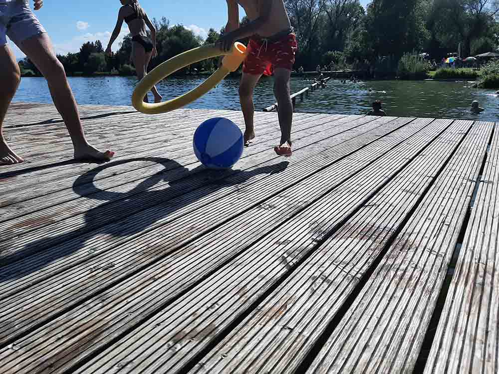 zwei Kinder in Badehosen spielen auf einem Holzsteg am See mit einem Wasserball und einer Schwimmnudel